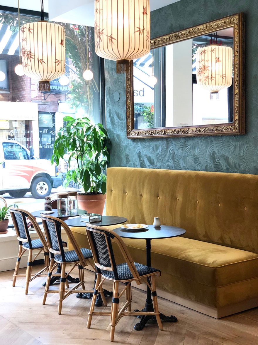 cafe chairs and gold velvet bench with round tables in front of window with plant and mirror