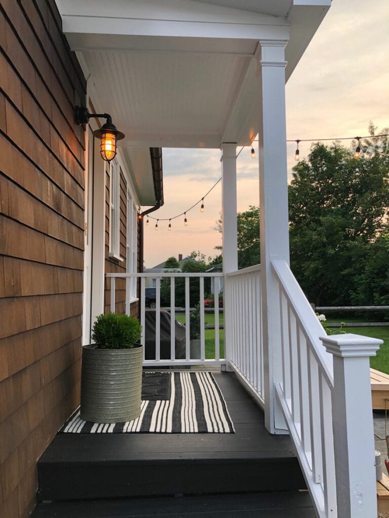 side porch, light, planter, boxwood, striped rug, railing painted white, sky with trees