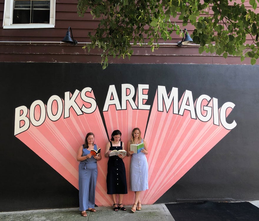 3 girls in front of wall mural on street with books in hand