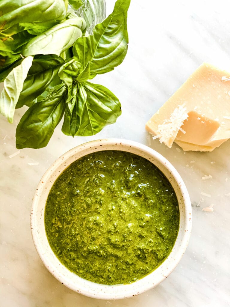 A bowl of freshly made basil pesto with basil leaves and cheese next to it.