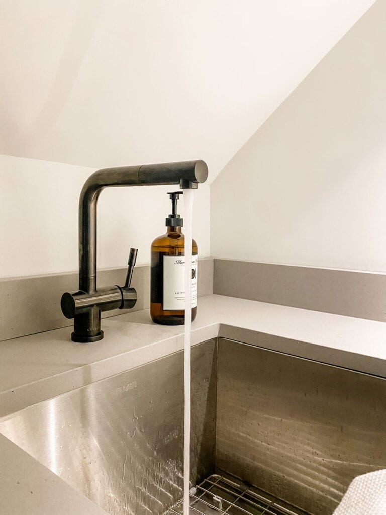 A stainless steel sink in a new laundry room with a simple black faucet with water running. An amber bottle of soap is next to the faucet.