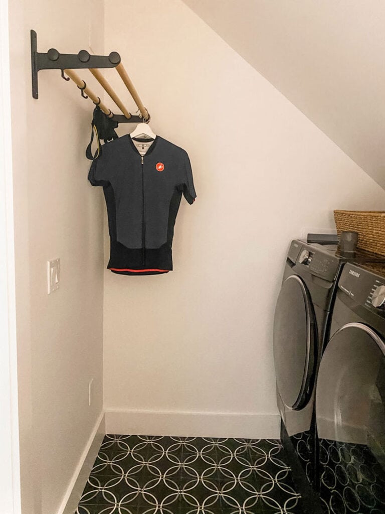 A utility/drying rack hangs on the wall next to a black washer and dryer in a newly renovated laundry room. The floor tile is a black, white, and gray design.