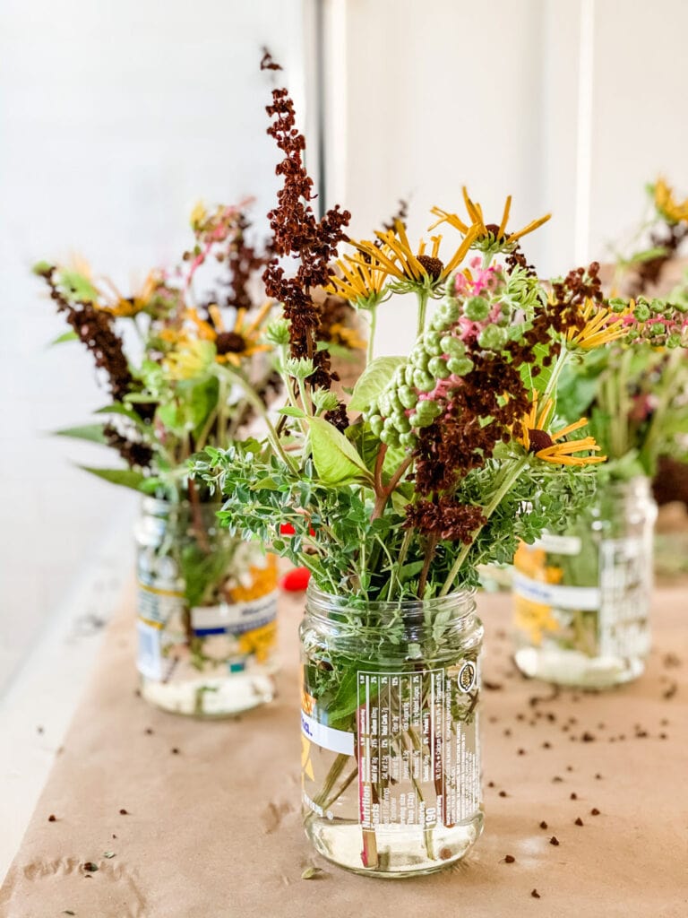 Glass jars of foraged flowers and herbs sitting on Kraft paper on stainless kitchen island.