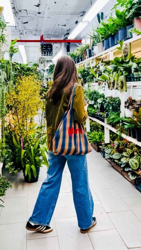 Annie Diamond of mostlovelythings.com with a Merci tote bag over her shoulder is walking through a plant store at the New York Flower market.