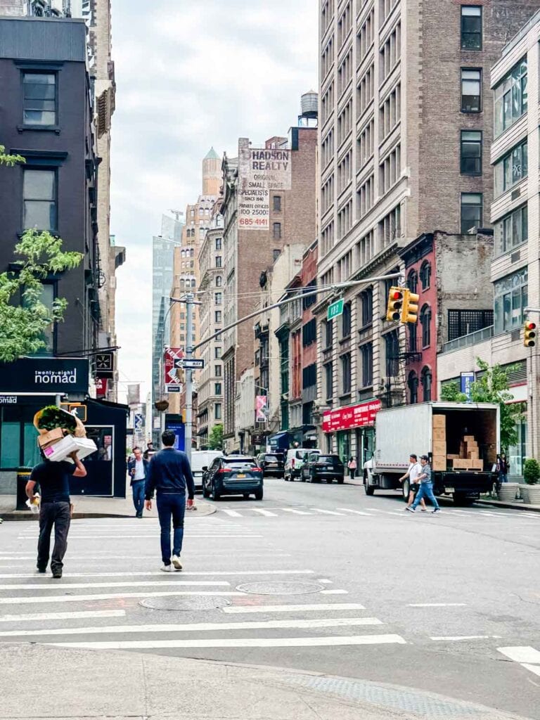 A man crossing the street in New York City with a large box and large brown paper bag of flowers on his shoulders.