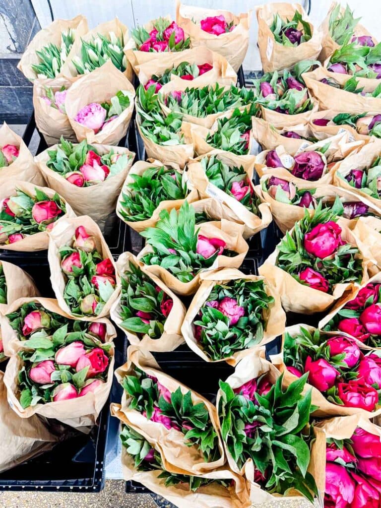 Different shades of peony flowers wrapped in brown paper are stacked in front of this storefront.