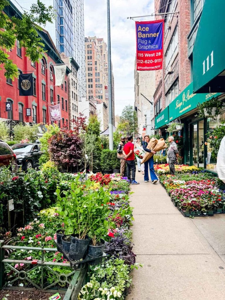 A woman at the NYC Flower Market is carrying large bundles of flowers wrapped in brown paper.
