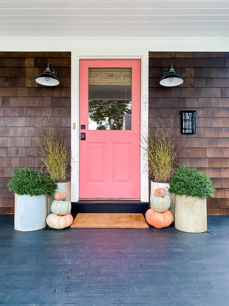 A pink front door with black porch lights on each side, a black mailbox, several larges pots planted with boxwoods and sea grass and stacked pumpkins next to them