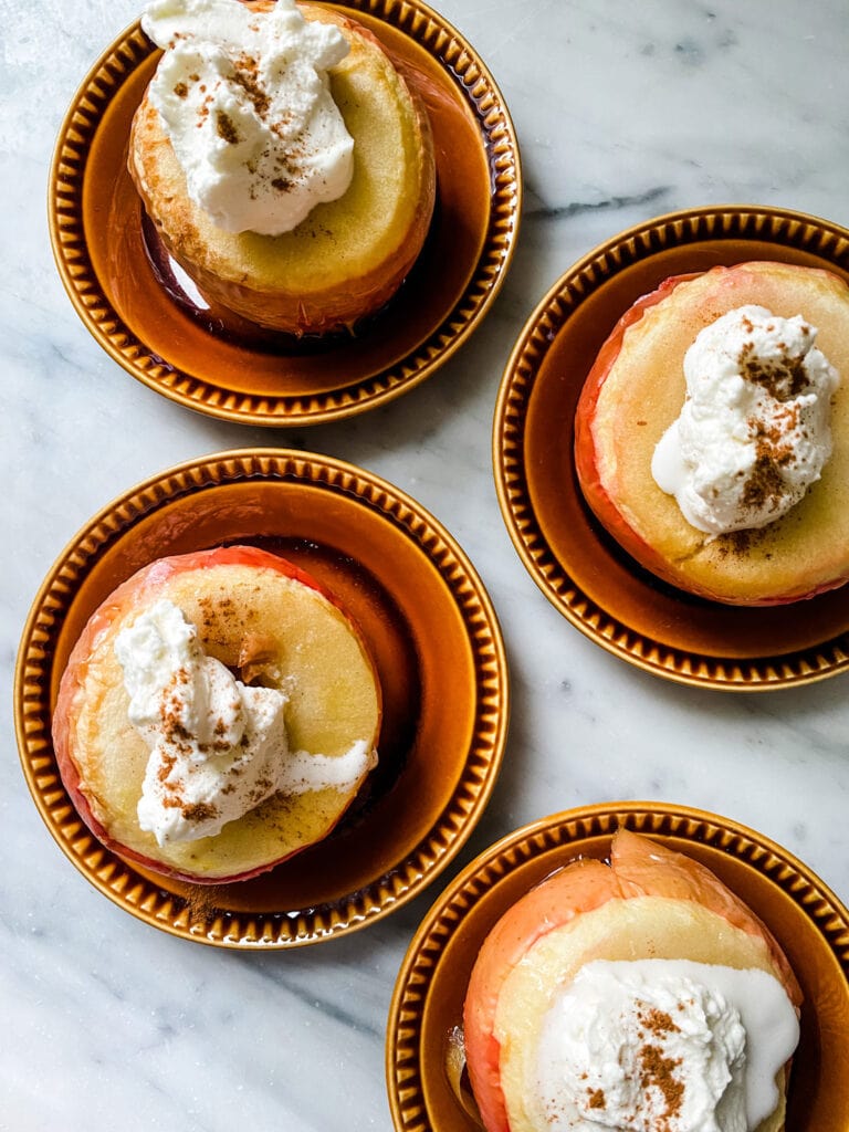 Cinnamon Baked Apples with a dollop of cream sprinkled with cinnamon on small amber colored mini plates on marble countertop