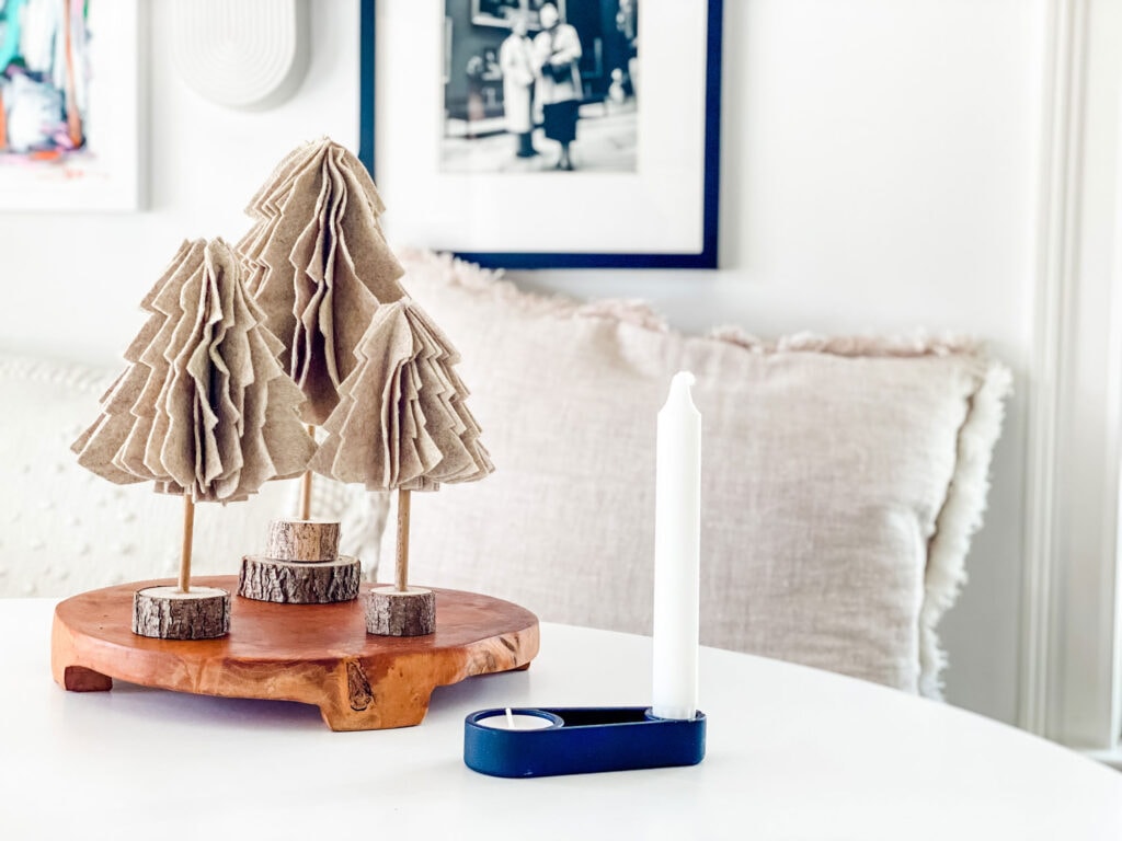 Scandinavian felt Christmas trees are displayed on wooden trivet on a white table. Next to it is a black candle holder from Copenhagen with a votive and simple white stick candle.
