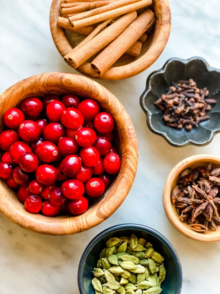 Cinnamon sticks, cloves, cardamom pods, star anise, orange, and cranberries in various bowls.