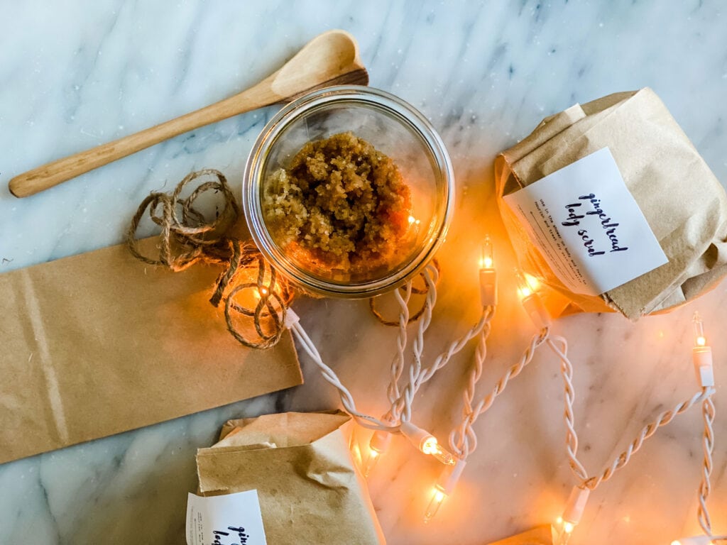Make this easy gingerbread body scrub. Scrub is in a glass jar next to a wooden mixing spoon. Brow bags and a sticker are used for packaging as a gift.