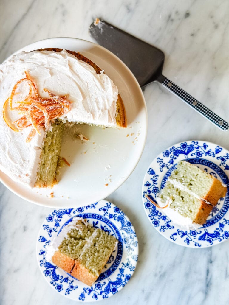 Blood orange olive oil cake is on a small white cake stand and next to the cake stand are two slices of cake on small blue and white plates.