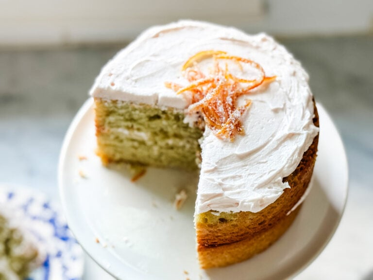 A blood orange olive oil decorated with butter cream frosting and candied orange zest is on a white cake stand.