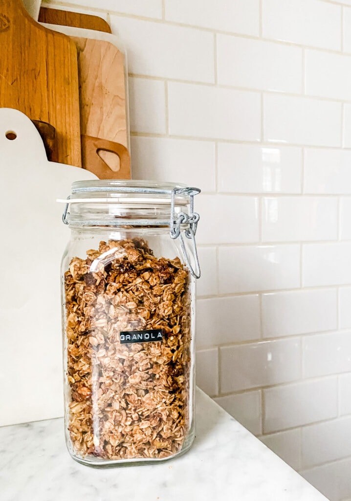 granola in a glass jar in a kitchen with cutting boards against a wall with subway tile