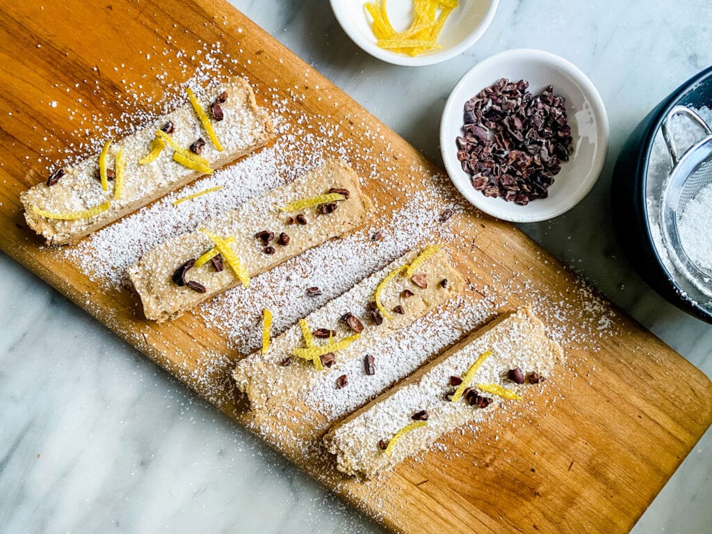 Twice-baked shortbread topped with chocolate chips and lemon rind, and dusted with confectioner's sugar is on a long, wooden cutting board.