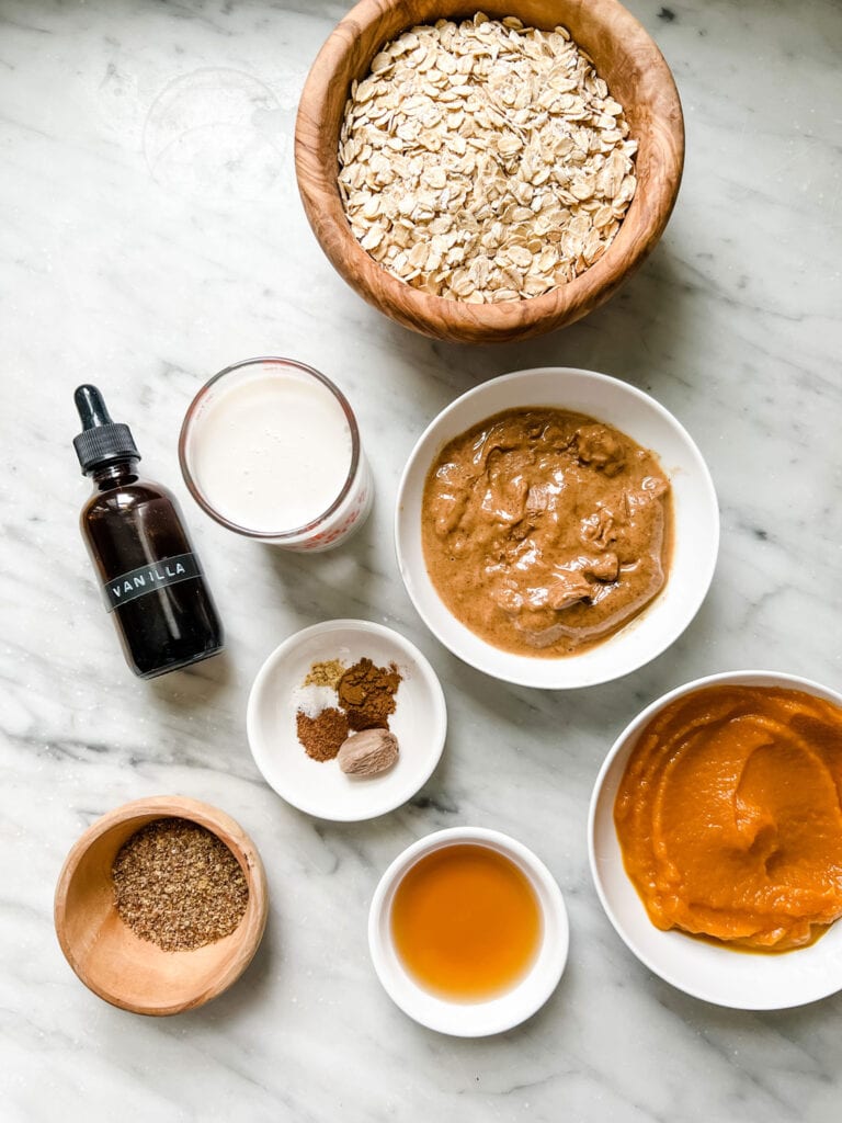 Ingredients for pumpkin pie overnight oats are on a countertop in small white bowls with a bottle of vanilla extract and a glass of milk.