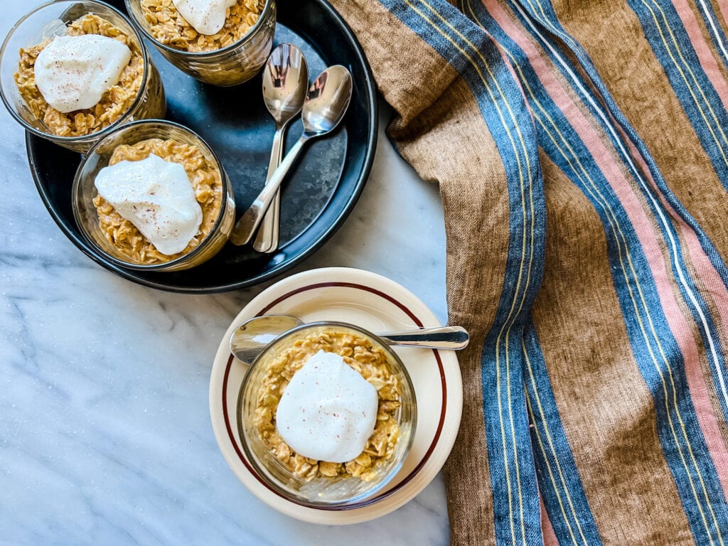 Four servings of pumpkin pie overnight oats in small glass containers. One is resting on a brown plate with a spoon. Three are on a black serving tray with spoons. A Lebico Linen tea towel is draped along the side.