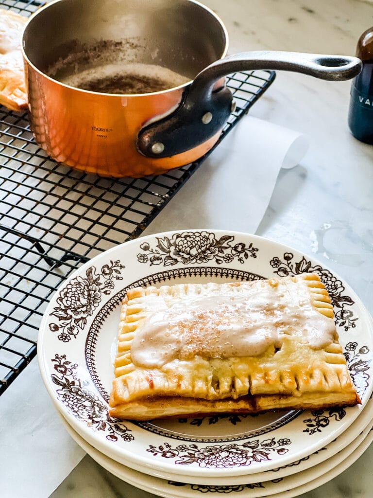 A homemade apple pie pop tart is on a small white plate with a brown floral design. Next to the plate is a wire cooling rack and a copper pot that was used for making the glaze.