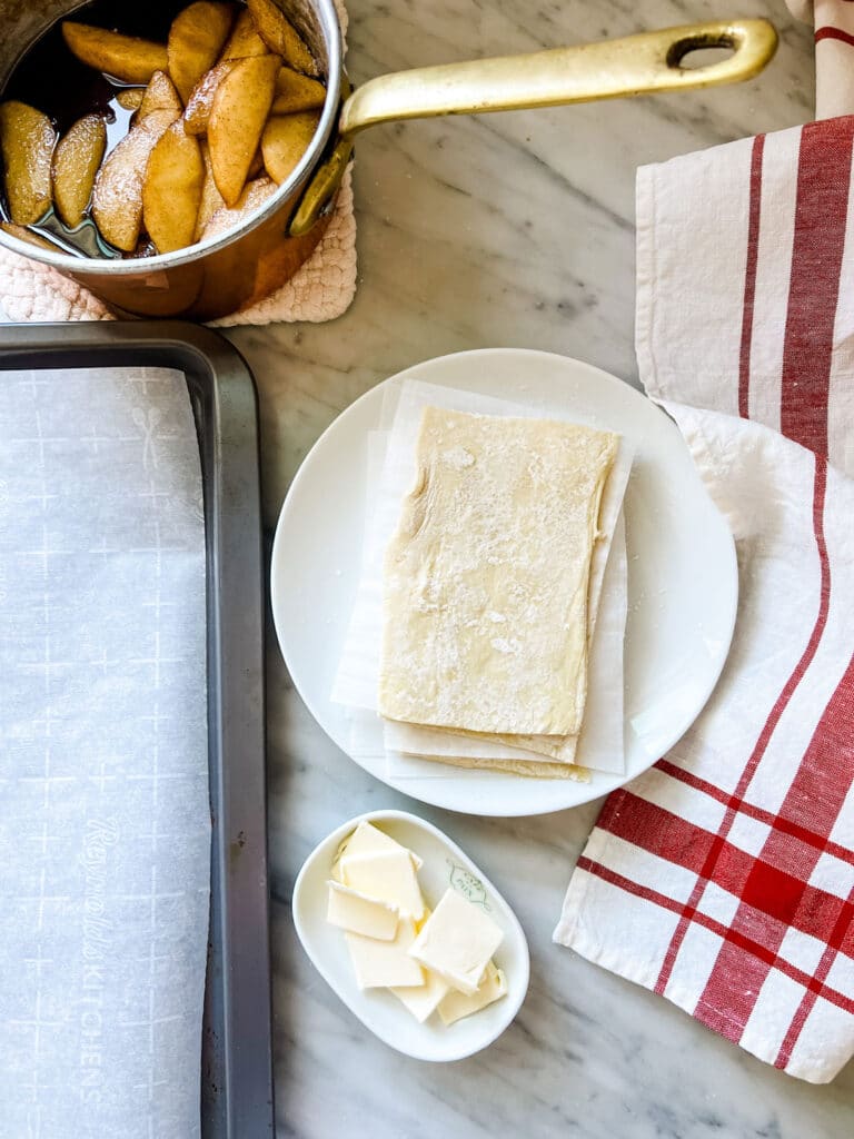 puff pastry rectangles on plate, baking sheet with parchment paper, red and white linen towel, copper pot with cooked apples 