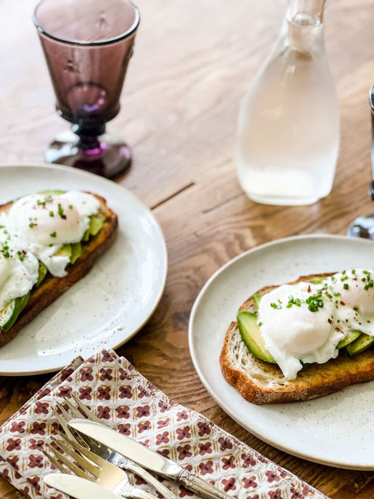 Simple Avocado & Poached Egg Tartine on plates, with printed linen napkins, silverware and bottle of water on rustic wood table