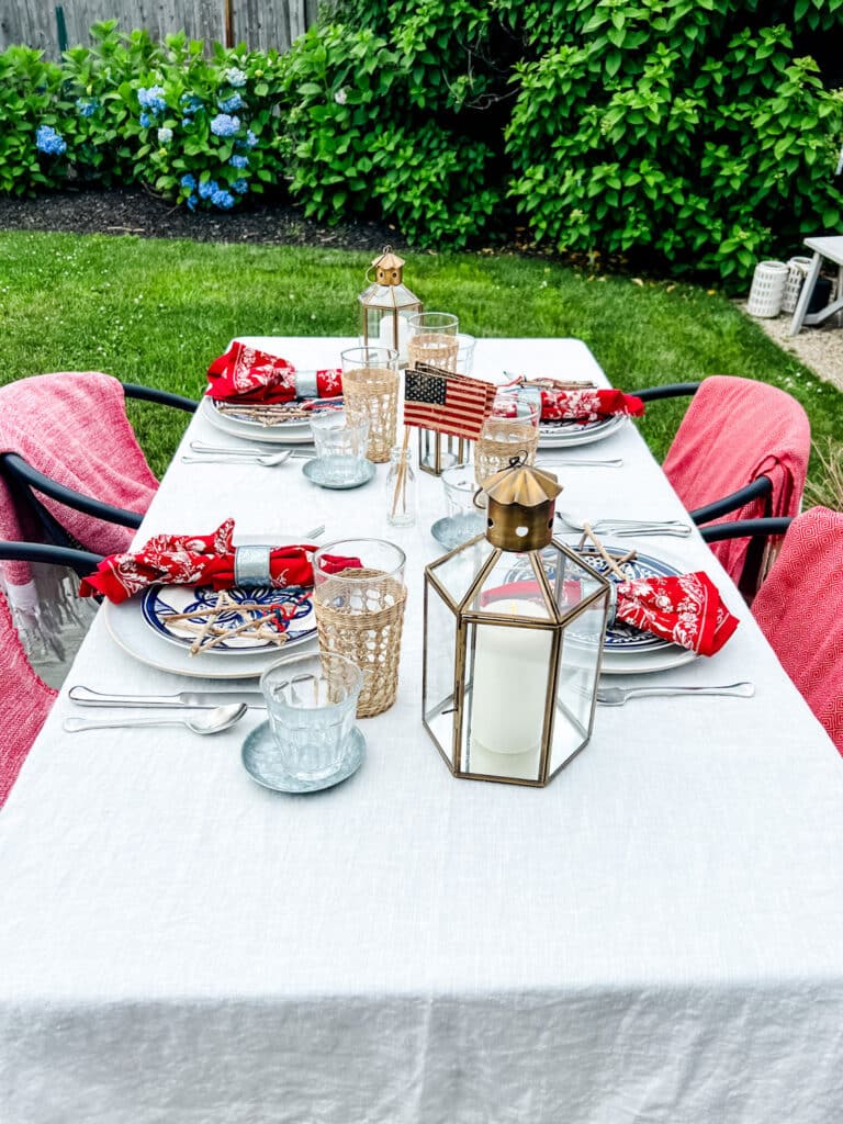 table with lanterns, plates, red print napkins, red towels draped on chairs, yard in background during summer 