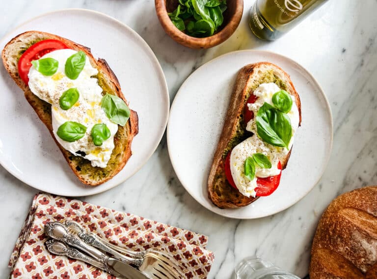 Caprese tartines are served on small white plates with light pink rims. Next to the plates are linen napkins, cutlery, a bowl of freshly picked basil, and a bottle of good extra-virgin olive oil.