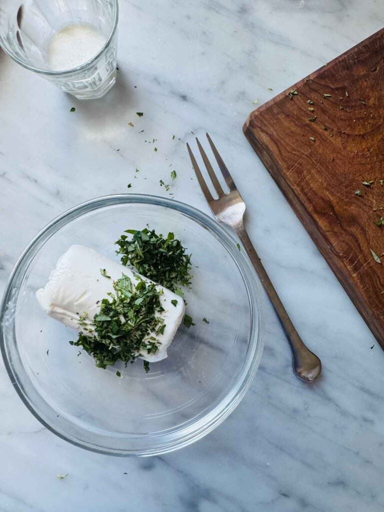 Goat cheese is in a small glass bowl with with fresh herbs and is ready for mixing.
