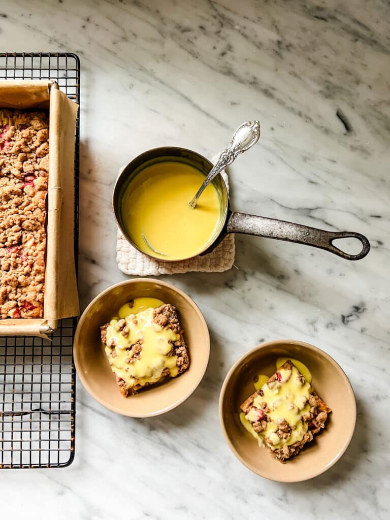 2 small bowls with rhubarb barsa with vanilla cream sauce on top, pan of sauce and bars The Best Rhubarb Crumb Bar Recipe With Vanilla Cream