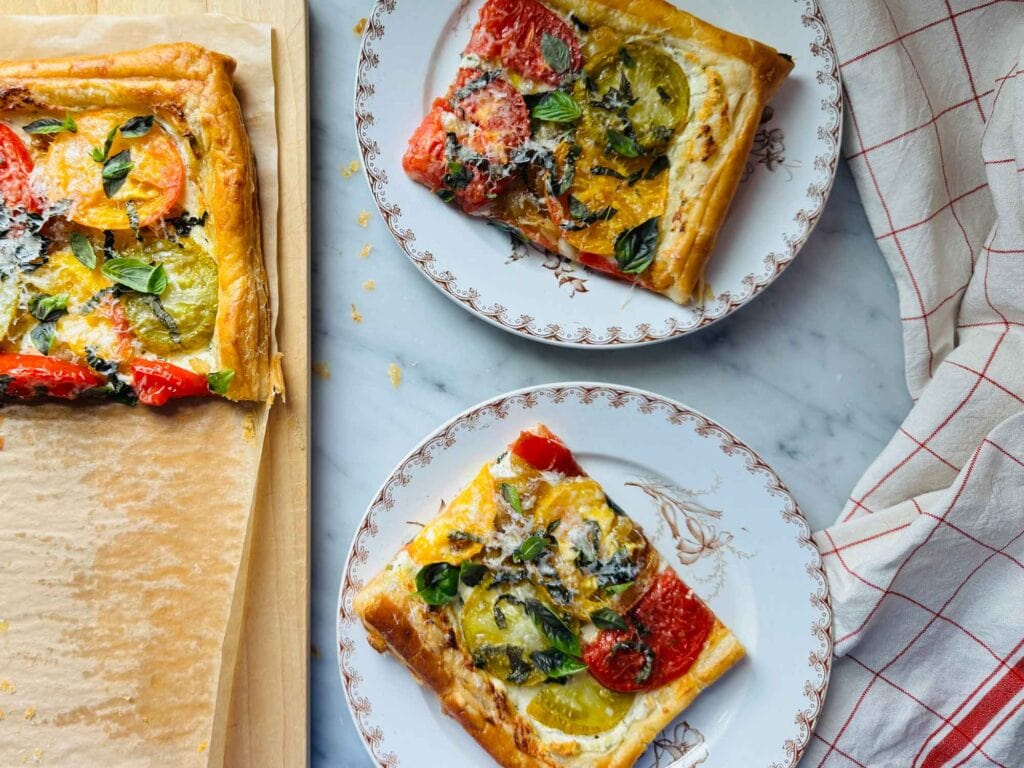 A puff pastry tart is served on small vintage French lunch plates and is sitting next to the remaining tart on a wooden cutting board.