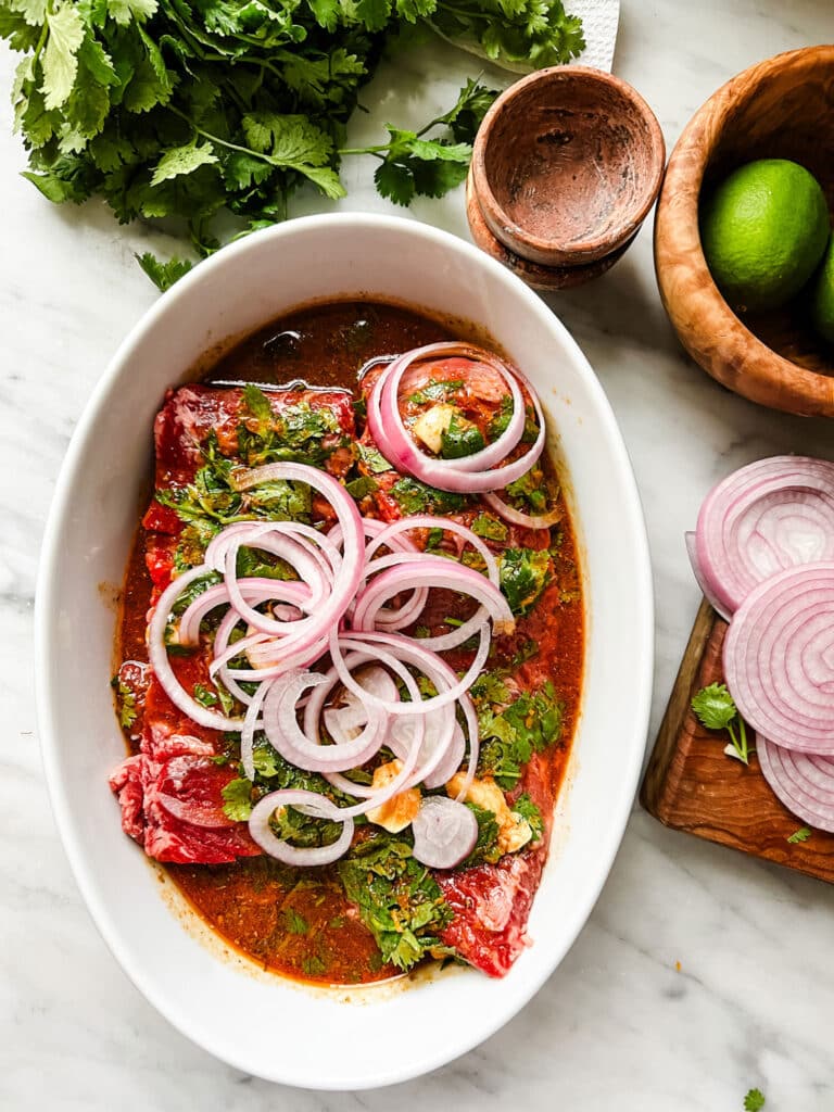 Skirt steak marinating in a shallow dish, sliced onions on a cutting board. Limes in a wooden bowl and a bunch of cilantro.
