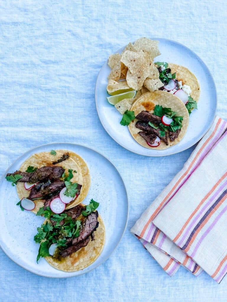 Authentic carne asada street tacos on individual plates, garnished with radishes, cilantro, tomatillo salsa, and lime wedges with tortilla chips on the side
