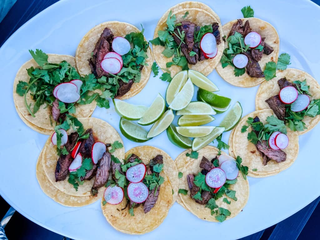 Carne Asada street tacos on white platter and garnished with thin slices of radish and cilantro. Lime wedges in center of platter.