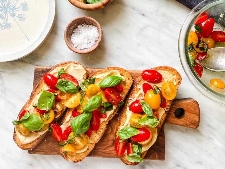 Healthy Mediterranean hummus, basil, and tomato tartines are prepared and resting on a wooden cutting board. A dish of coarse sea salt is next to the cutting board.