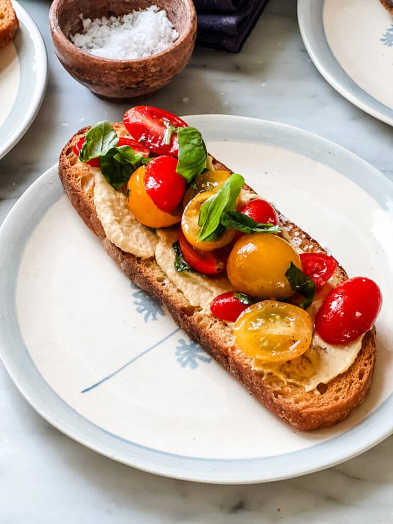 Single plate with piece of sliced bread with hummus spread topped with fresh tomatoes and basil.