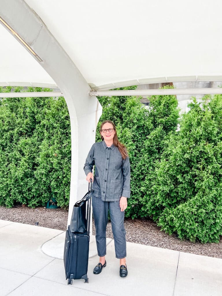 Older woman wearing suit and strnding by a suitcase in front of evergreen trees