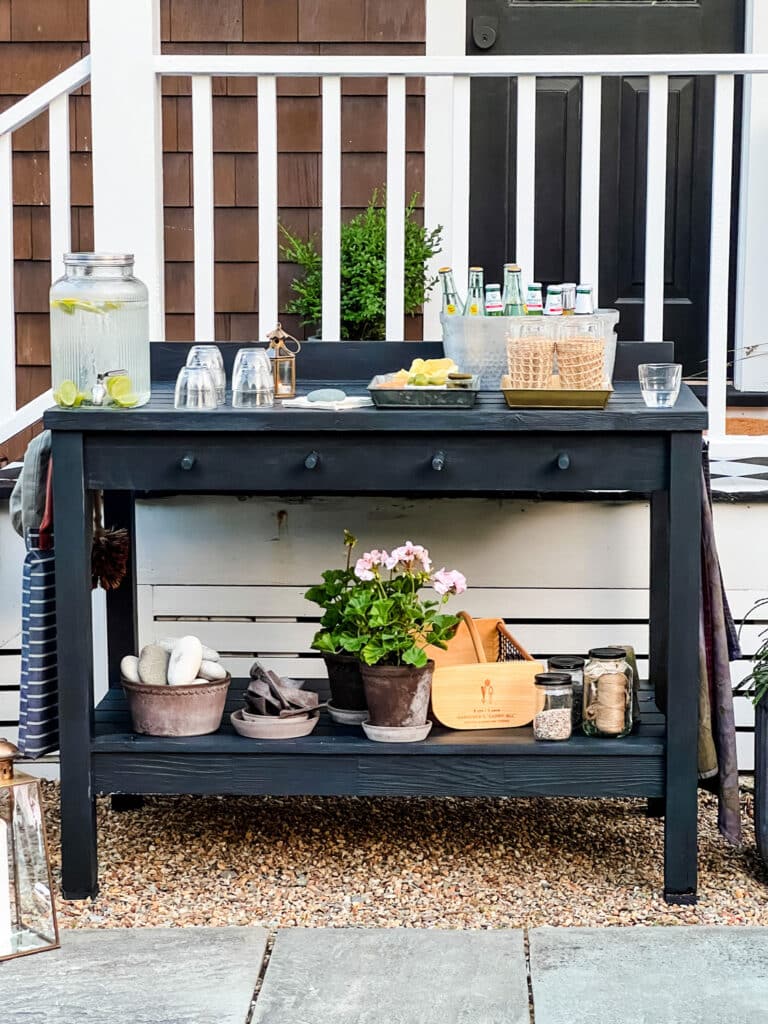 Black potting bench with garden tools and supplies on lower shelf, and summer entertaining items on top