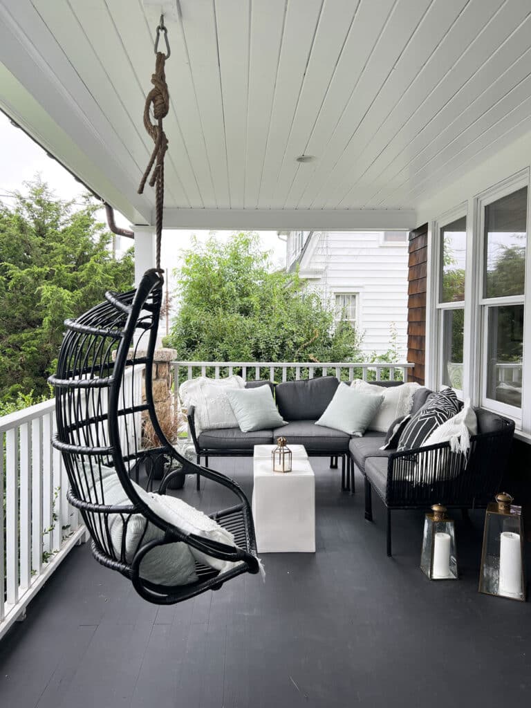 Hanging black rattan wicker chair on a porch with sofa and pillow and black wood floors. Lanterns with candles sitting on the floor.