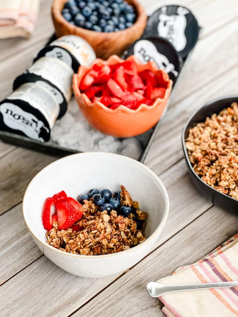 White bowl of granola and fresh fruit in the foreground. In the background is a tray of ice with yogurt and fresh strawberries, blueberries next to a black serving bowl of granola.