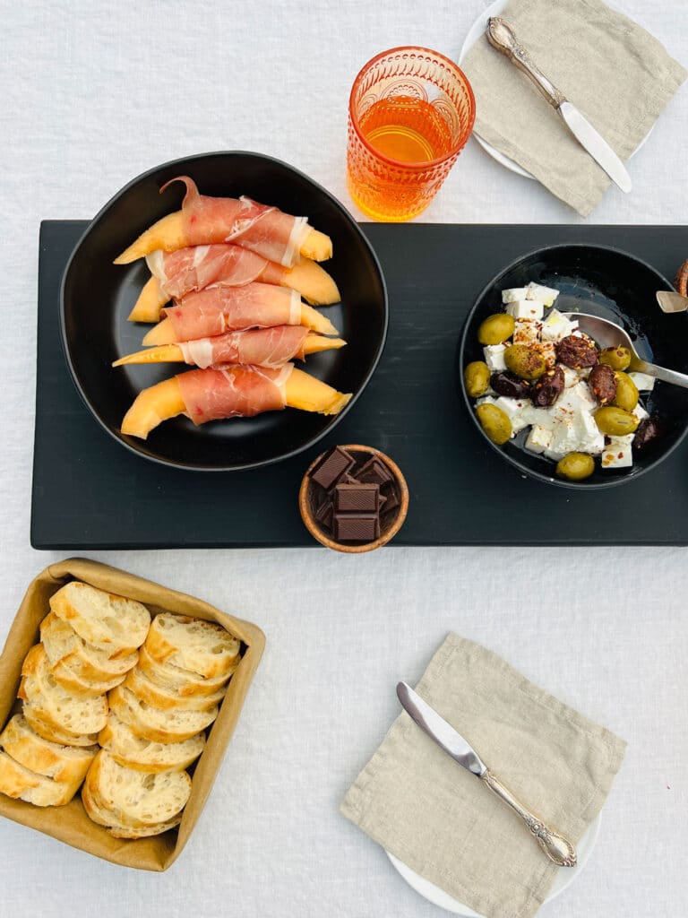 Black farmtable plank on white tablecloth with cantaloupe wrapped in prosciutto withe crackers in a basket