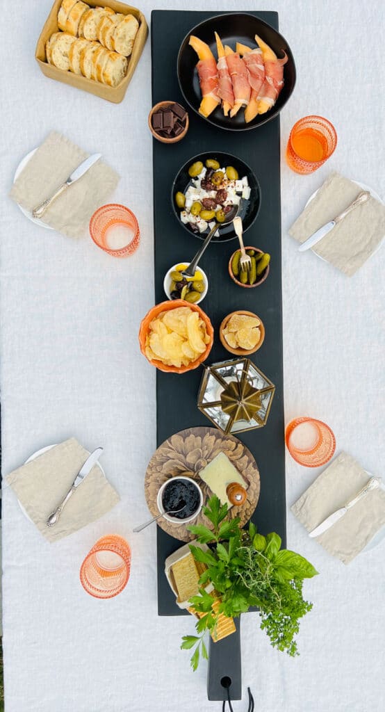 Long black farmtable plank cheese and charcuterie board on white table cloth with a table setting for four people