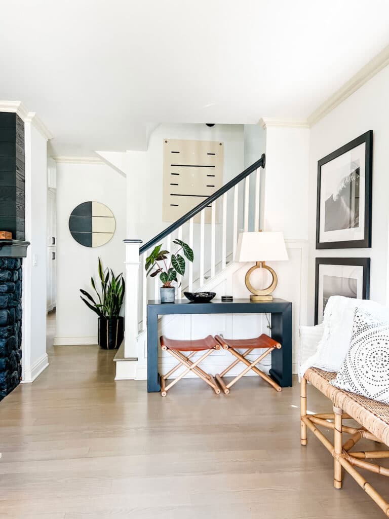 Console table with stools under staircase, canvas painting hanging on wall in staircase area and round wood black and white art hanging on wall.
