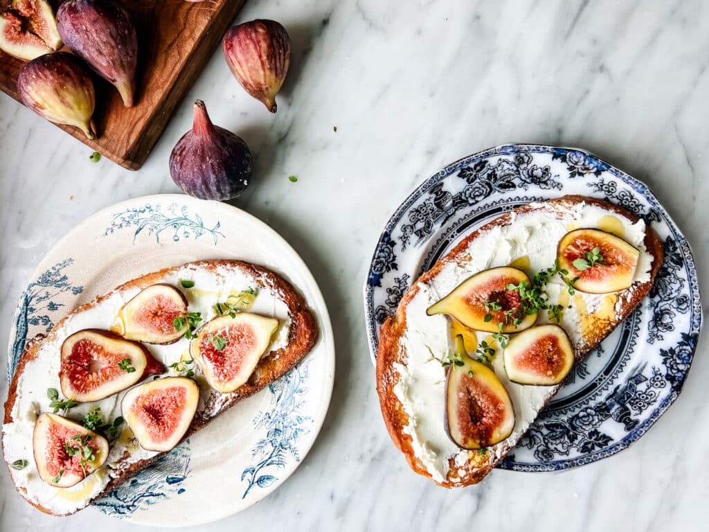Two plates sitting on a marble countertop with toasted bread topped with goat cheese, fresh figs, and thyme.
