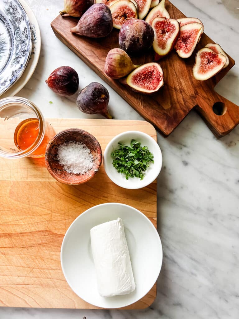 Two wood cutting boards sitting on a marble countertop. One cutting board has fresh sliced figs while the second contains honey, sal, thyme and goat cheese