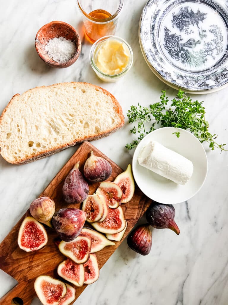 Freshly sliced figs, a bowl of goat cheese, thyme, bread salt, butter, and honey sitting on a marble countertop