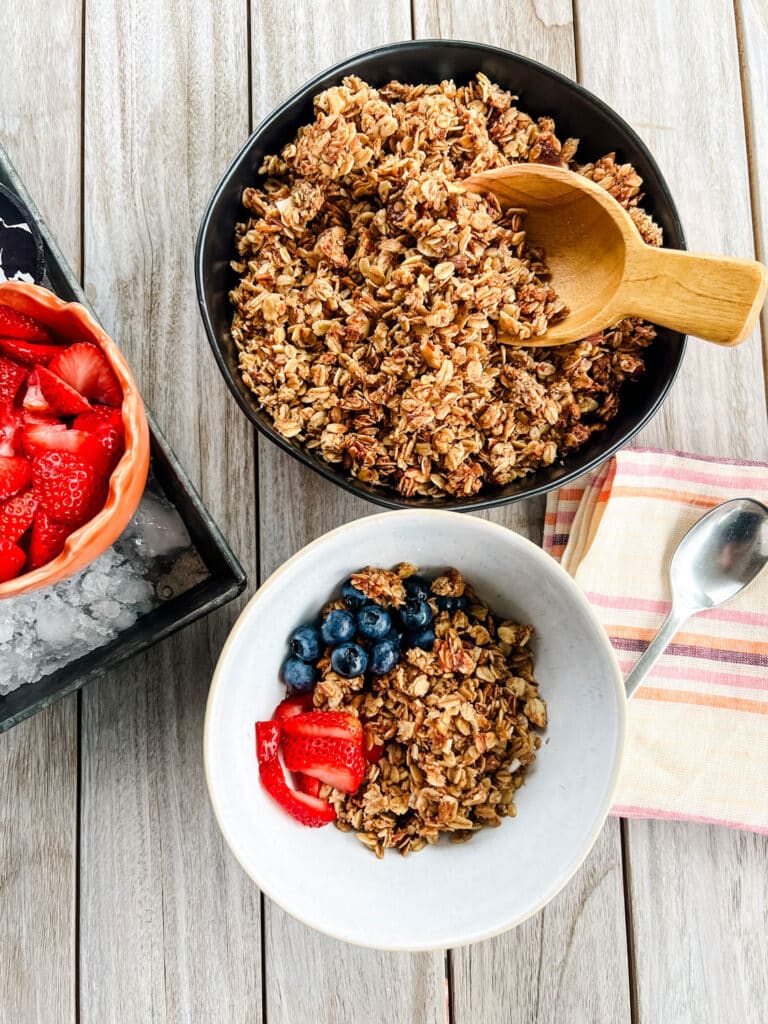 White bowl full of granola and fresh strawberries and blueberries next to a black serving bowl with a wooden serving scoop Easy Homemade Granola Recipe That's Healthy and Delicious