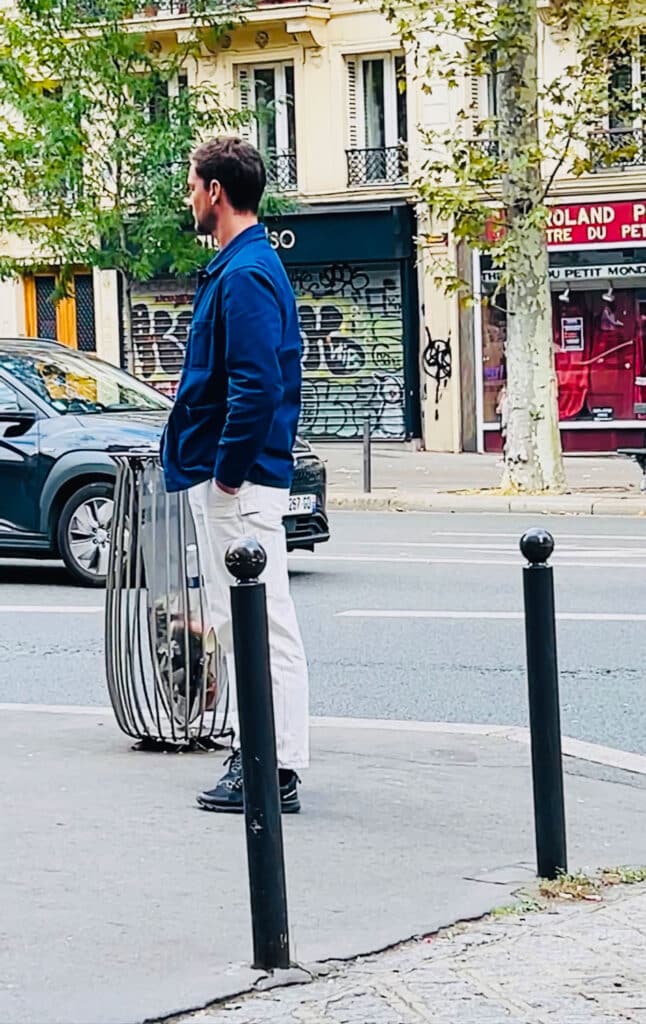 A young man standing on a street corner in Paris wearing a blue French chore coat.