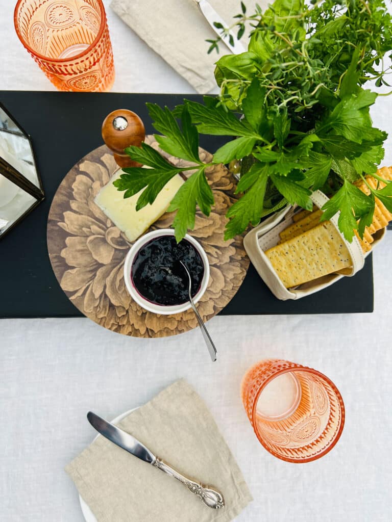 Table seeting with cheese, crackers and fresh herbs on black farmtable plank cheese and charcuterie board with plates and orange drinking glasses