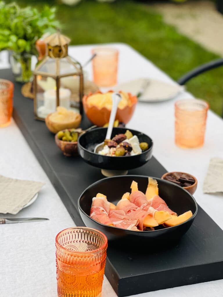 Outdoor table with a white tablecloth, dinnerware setting for four people and a black farmtable plank cheese and charcuterie board