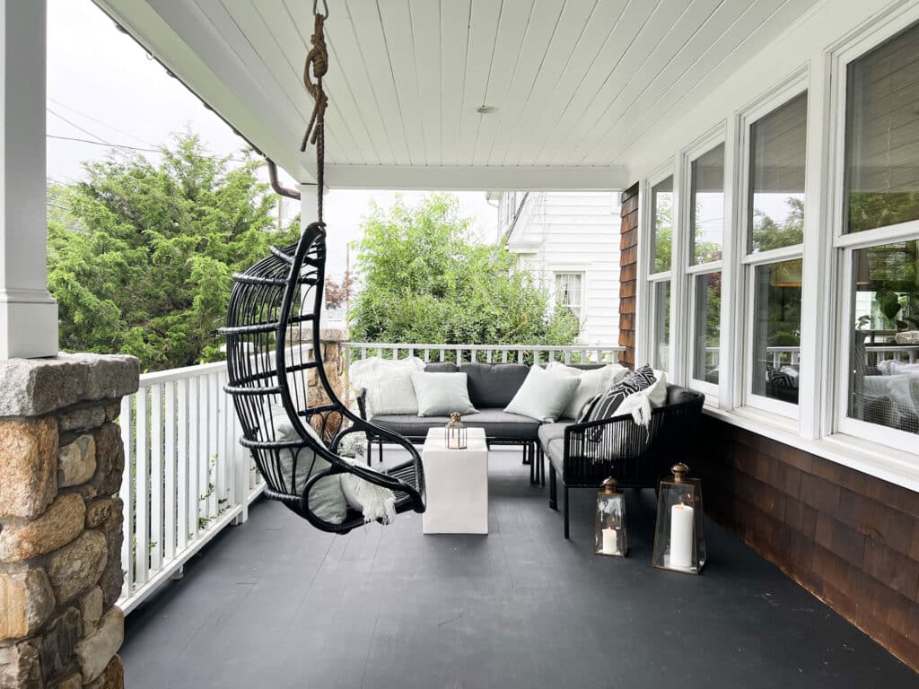 Hanging black rattan wicker chair on a porch with sofa and pillow and black wood floors. Lanterns with candles sitting on the floor.